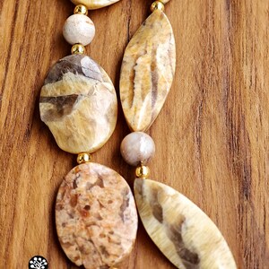 Puede incluir: Un collar con piedras pulidas de diversas formas en tonos beige, marr&oacute;n y ocre, intercaladas con peque&ntilde;as cuentas redondas de metal color oro. Las piedras presentan vetas naturales y texturas variadas dispuestas en un patr&oacute;n lineal.