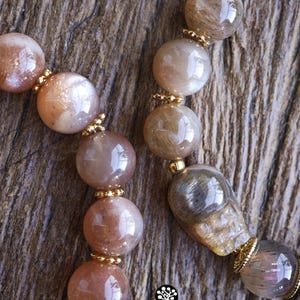 May include: Two beaded bracelets featuring polished, translucent, and opaque round beads in shades of peach, tan, and grey. The beads are interspersed with small, gold-toned metal spacers. One bracelet includes a carved skull-shaped bead.