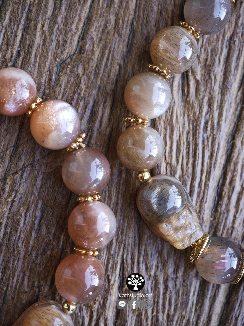 May include: Two beaded bracelets featuring polished, translucent, and opaque round beads in shades of peach, tan, and grey. The beads are interspersed with small, gold-toned metal spacers. One bracelet includes a carved skull-shaped bead.
