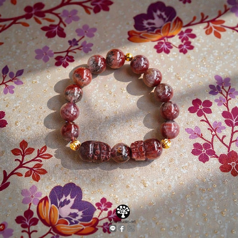 Peut inclure: Un bracelet de perles compos&eacute; de perles en pierre gemme polies, de couleur rouge et sombre, altern&eacute;es avec deux perles rectangulaires sculpt&eacute;es et de petites entretoises en m&eacute;tal dor&eacute; textur&eacute;, dispos&eacute;es en cercle sur une surface claire.