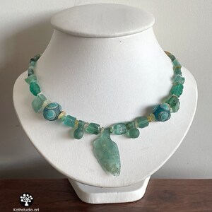 Puede incluir: Collar artesanal con cuentas de vidrio verde mar y un colgante central. El collar incluye cuentas decorativas con dise&ntilde;os azules y blancos y se exhibe sobre un busto blanco. Una pieza de joyer&iacute;a &uacute;nica.