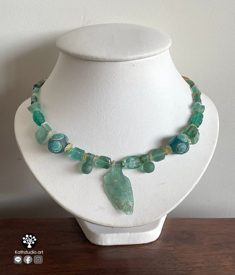 Puede incluir: Collar artesanal con cuentas de vidrio verde mar y un colgante central. El collar incluye cuentas decorativas con dise&ntilde;os azules y blancos y se exhibe sobre un busto blanco. Una pieza de joyer&iacute;a &uacute;nica.