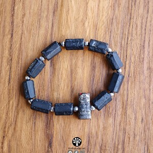 May include: A stretch bracelet featuring cylindrical black tourmaline beads alternating with small metallic silver-toned spacer beads. A single carved gray stone charm depicting a small creature with red-painted ears is integrated into the design.