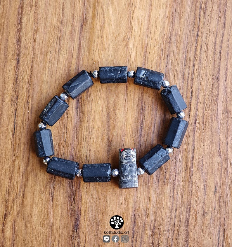 May include: A stretch bracelet featuring cylindrical black tourmaline beads alternating with small metallic silver-toned spacer beads. A single carved gray stone charm depicting a small creature with red-painted ears is integrated into the design.