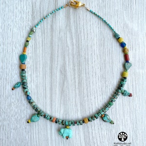 Op de afbeelding: Een turquoise kralenketting met een gouden sluiting. De ketting heeft verschillende kleurrijke kralen, waaronder turquoise, geel, rood en blauw. Het heeft kleine vogel- en olifantenbedels. De ketting wordt gepresenteerd op een licht houten oppervlak.