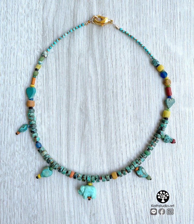 Op de afbeelding: Een turquoise kralenketting met een gouden sluiting. De ketting heeft verschillende kleurrijke kralen, waaronder turquoise, geel, rood en blauw. Het heeft kleine vogel- en olifantenbedels. De ketting wordt gepresenteerd op een licht houten oppervlak.