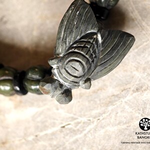 Puede incluir: Una pulsera de piedra verde oscuro con un dije de cigarra tallado como pieza central. La pulsera consta de cuentas verdes redondeadas y texturizadas intercaladas con cuentas espaciadoras negras m&aacute;s peque&ntilde;as y lisas. El logo KATHSTUDIO BANGKOK es visible.