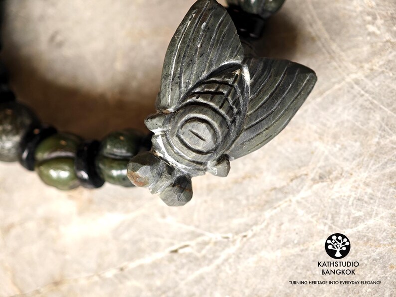 Puede incluir: Una pulsera de piedra verde oscuro con un dije de cigarra tallado como pieza central. La pulsera consta de cuentas verdes redondeadas y texturizadas intercaladas con cuentas espaciadoras negras m&aacute;s peque&ntilde;as y lisas. El logo KATHSTUDIO BANGKOK es visible.
