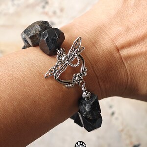 May include: A bracelet composed of large, faceted, matte black beads strung together. The piece features a prominent silver-toned metal toggle clasp shaped like a dragonfly with intricate filigree wings and a small flower detail at the base.