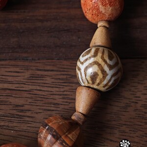 May include: Close-up of a handcrafted necklace featuring wooden beads in various shapes and sizes. The necklace includes orange, spherical beads with a porous texture, and patterned, spherical beads with brown and white designs. The necklace is displayed on a wooden surface.