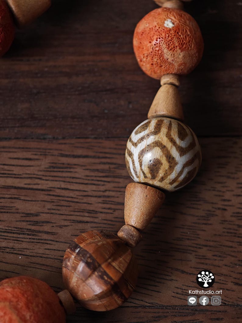 May include: Close-up of a handcrafted necklace featuring wooden beads in various shapes and sizes. The necklace includes orange, spherical beads with a porous texture, and patterned, spherical beads with brown and white designs. The necklace is displayed on a wooden surface.