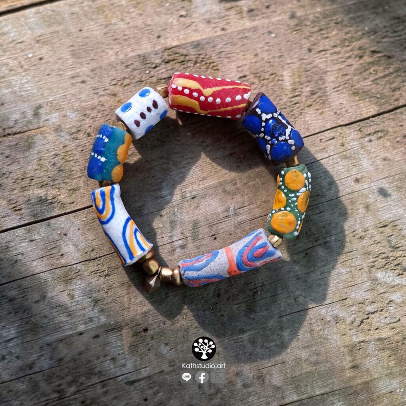 May include: A bracelet composed of cylindrical beads featuring hand-painted patterns in red, blue, yellow, white, and green, interspersed with small metallic spacer beads. The beads display various geometric designs including dots and swirls.