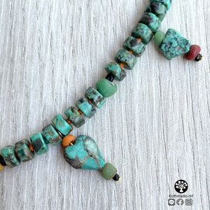 Op de afbeelding: Close-up van een turquoise kralenketting met een hartvormige hanger. De ketting heeft vierkante en onregelmatige kralen in tinten turquoise, groen en bruin. Kleine goudkleurige accenten en zwarte kralen zijn ook zichtbaar. De ketting wordt gepresenteerd op een wit houten oppervlak.