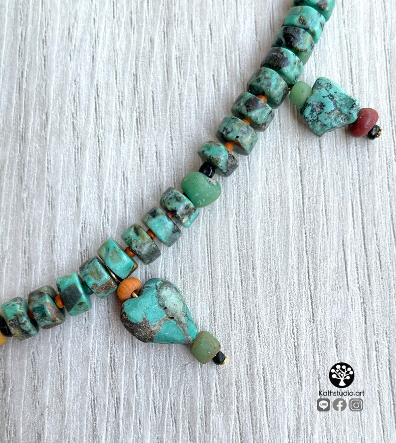 Op de afbeelding: Close-up van een turquoise kralenketting met een hartvormige hanger. De ketting heeft vierkante en onregelmatige kralen in tinten turquoise, groen en bruin. Kleine goudkleurige accenten en zwarte kralen zijn ook zichtbaar. De ketting wordt gepresenteerd op een wit houten oppervlak.