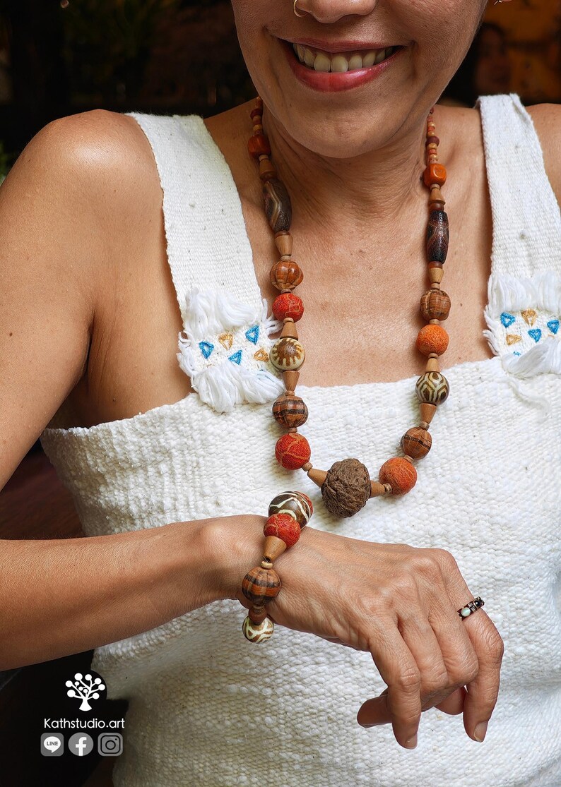 May include: A handcrafted necklace and bracelet set featuring a variety of wooden and textured beads in shades of brown, orange, and red. The jewelry is displayed against a white background.