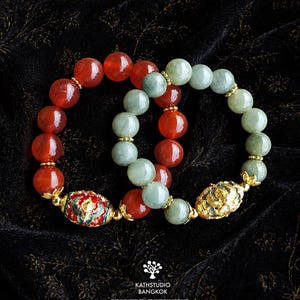 May include: Two beaded bracelets featuring polished red carnelian and pale green jade beads. One bracelet includes a gold-toned oval bead with red and green enamel floral patterns, while the other features a textured gold-toned oval bead.