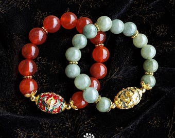May include: Two beaded bracelets featuring polished red carnelian and pale green jade beads. One bracelet includes a gold-toned oval bead with red and green enamel floral patterns, while the other features a textured gold-toned oval bead.