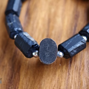 May include: A bracelet composed of elongated, rough-textured black stone beads alternating with small, faceted metallic silver beads. A central oval-shaped black bead features a small, etched numerical symbol on its flat surface.