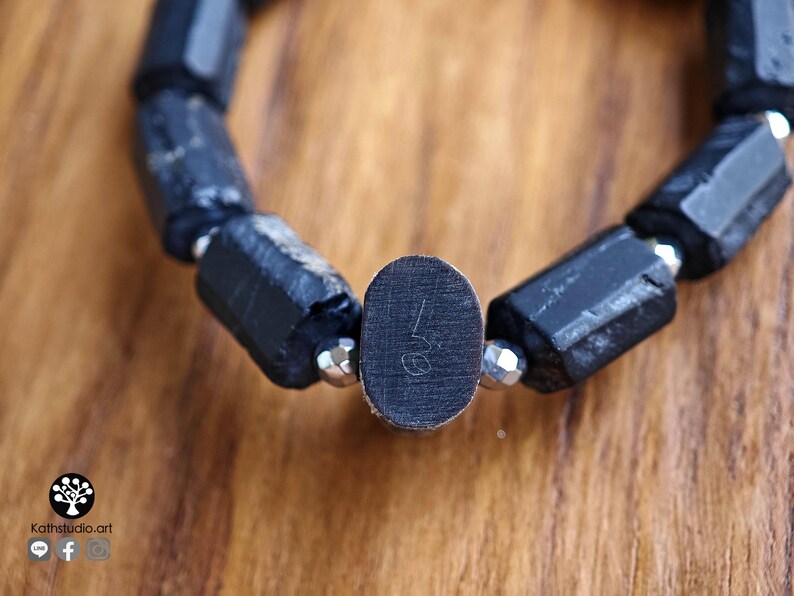 May include: A bracelet composed of elongated, rough-textured black stone beads alternating with small, faceted metallic silver beads. A central oval-shaped black bead features a small, etched numerical symbol on its flat surface.