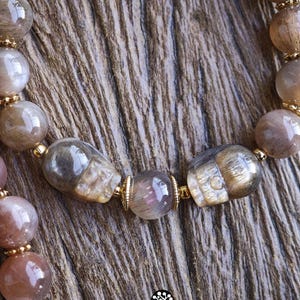 May include: A beaded bracelet featuring polished, translucent brown and grey rutilated quartz beads interspersed with two carved skull-shaped beads and small gold-toned metal spacers. The text Kathstudio.art is printed at the bottom of the image.