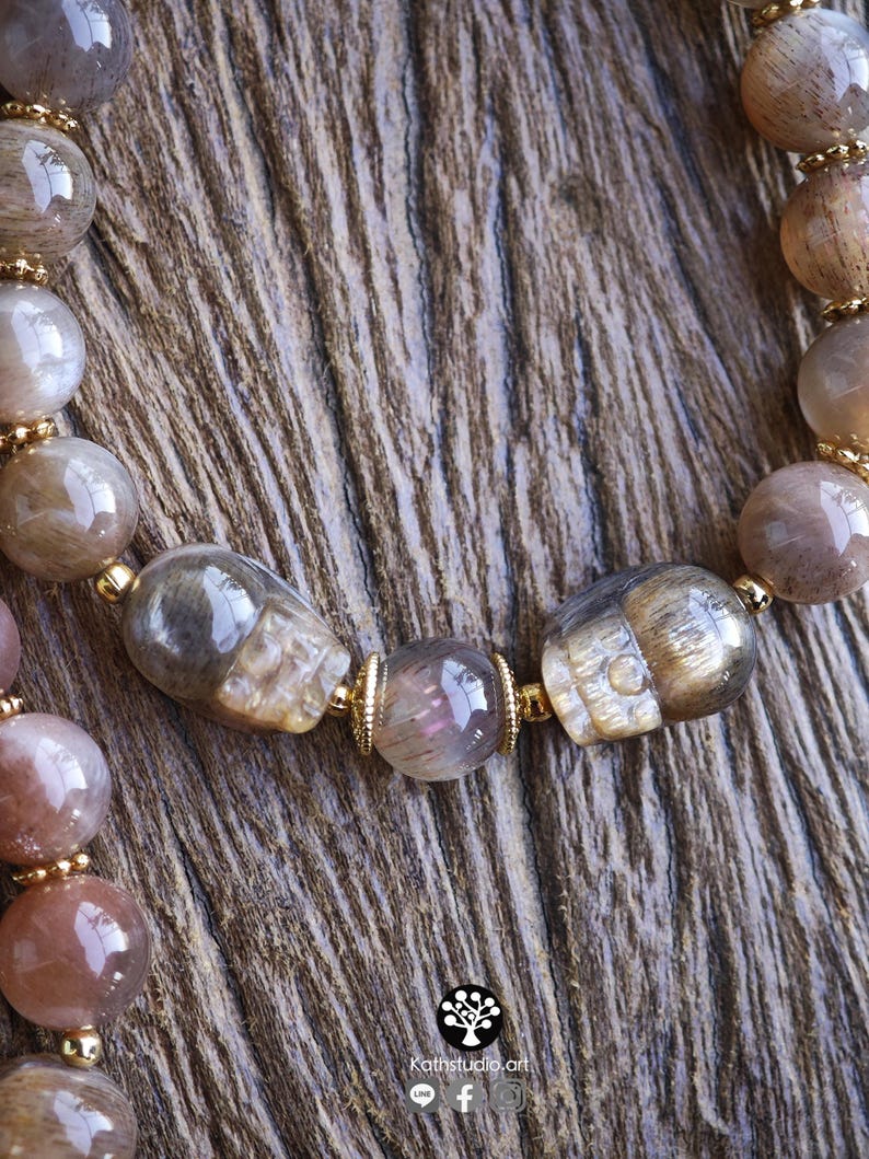 May include: A beaded bracelet featuring polished, translucent brown and grey rutilated quartz beads interspersed with two carved skull-shaped beads and small gold-toned metal spacers. The text Kathstudio.art is printed at the bottom of the image.