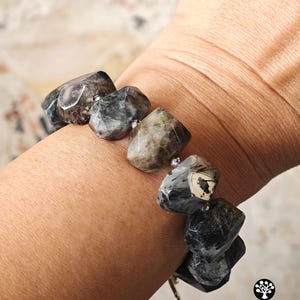 May include: A bracelet composed of large, faceted, irregular-shaped beads in shades of charcoal, slate gray, and earthy brown. The beads are separated by small, metallic silver-toned spacer beads, creating a textured, multi-faceted appearance.