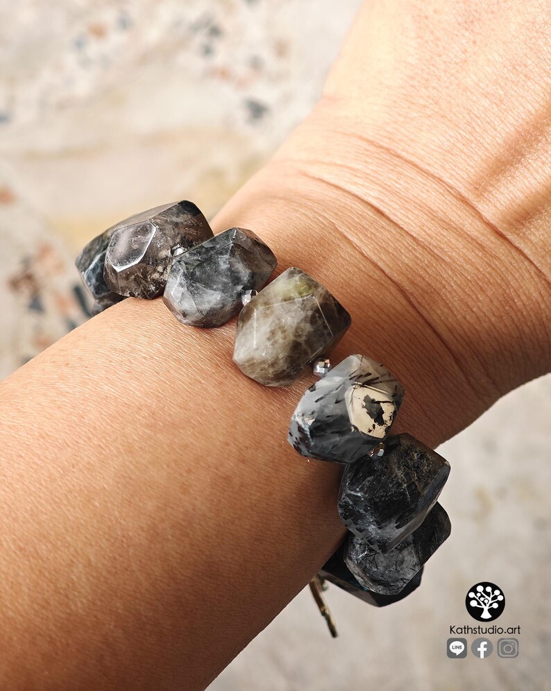 May include: A bracelet composed of large, faceted, irregular-shaped beads in shades of charcoal, slate gray, and earthy brown. The beads are separated by small, metallic silver-toned spacer beads, creating a textured, multi-faceted appearance.