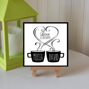 May include: Black and white illustration of two coffee mugs with steam rising from them. The mugs are labeled "Melissa" and "David" and the text "Love is Brewing" is above them.