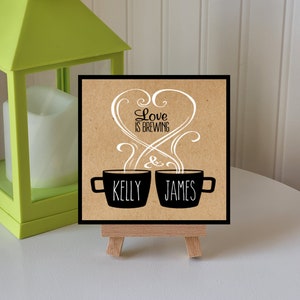 May include: A brown card with the text "Love is Brewing" in white cursive lettering. Two black coffee mugs with steam coming out of them are below the text. The mugs are labeled "Kelly" and "James". The card is on a small wooden easel.