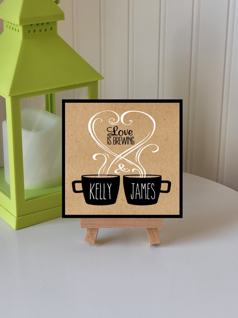 May include: A brown card with the text "Love is Brewing" in white cursive lettering. Two black coffee mugs with steam coming out of them are below the text. The mugs are labeled "Kelly" and "James". The card is on a small wooden easel.
