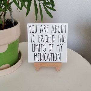 May include: White ceramic tile with black text that reads "You are about to exceed the limits of my medication." The tile is on a small wooden easel.