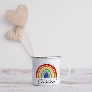 May include: White enamel mug with a rainbow design and the name "Cassie" printed on the bottom. The mug has a silver rim and handle.