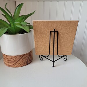 May include: A corkboard with a black metal easel stand. The corkboard is a light brown color and has a rough texture. The easel stand is made of thin metal and has a simple design.