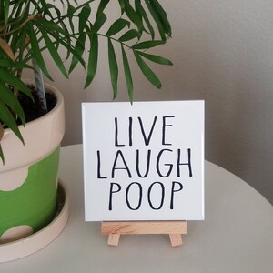 May include: White ceramic tile with black text that reads "Live Laugh Poop" on a small wooden easel. The tile is sitting on a white surface next to a potted green plant.