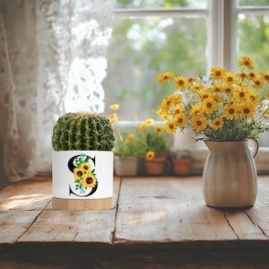 May include: White ceramic planter with a wooden base, featuring a sunflower design and the letter "S". A green cactus plant is inside the planter. The planter is on a wooden table with a bouquet of yellow flowers in a white vase.