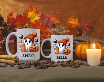 May include: Two white ceramic mugs with a Halloween theme. The mugs feature a cartoon ghost wearing a hat and holding a pumpkin. The mugs are personalized with the names "Amber" and "Bella".