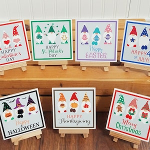 May include: Set of 8 small wooden easel stands with painted square signs featuring gnomes and holiday greetings. The signs include "Happy Valentine's Day", "Happy St. Patrick's Day", "Happy Easter", "Happy 4th of July", "Happy Halloween", "Happy Thanksgiving", and "Merry Christmas".