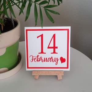 May include: White square tile with a red border and red text that says "14 February" with a red heart. The tile is on a small wooden easel.