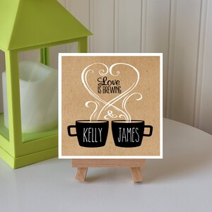 May include: A brown card with the text "Love is Brewing" in a white heart shape with swirls. Two black coffee mugs with steam coming out of them are below the heart. The mug on the left says "Kelly" and the mug on the right says "James".