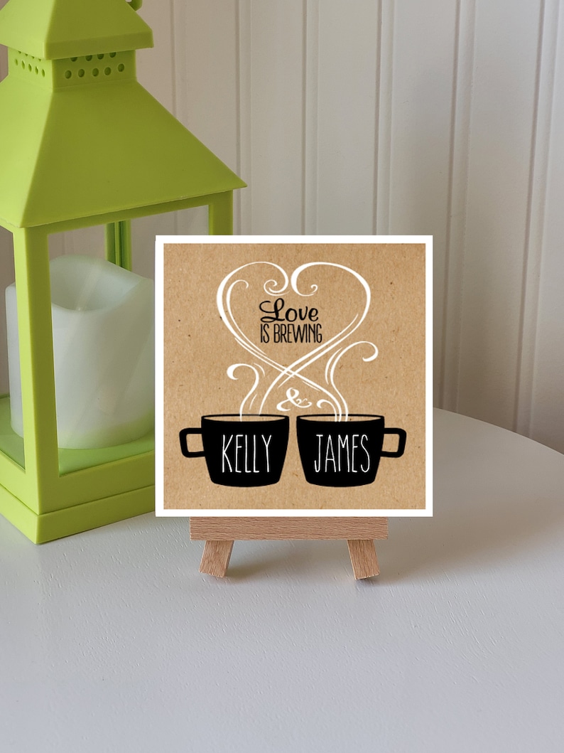 May include: A brown card with the text "Love is Brewing" in a white heart shape with swirls. Two black coffee mugs with steam coming out of them are below the heart. The mug on the left says "Kelly" and the mug on the right says "James".