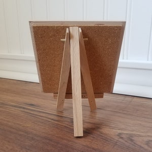 May include: A small wooden easel with a corkboard surface. The easel is made of natural wood and has a simple design.