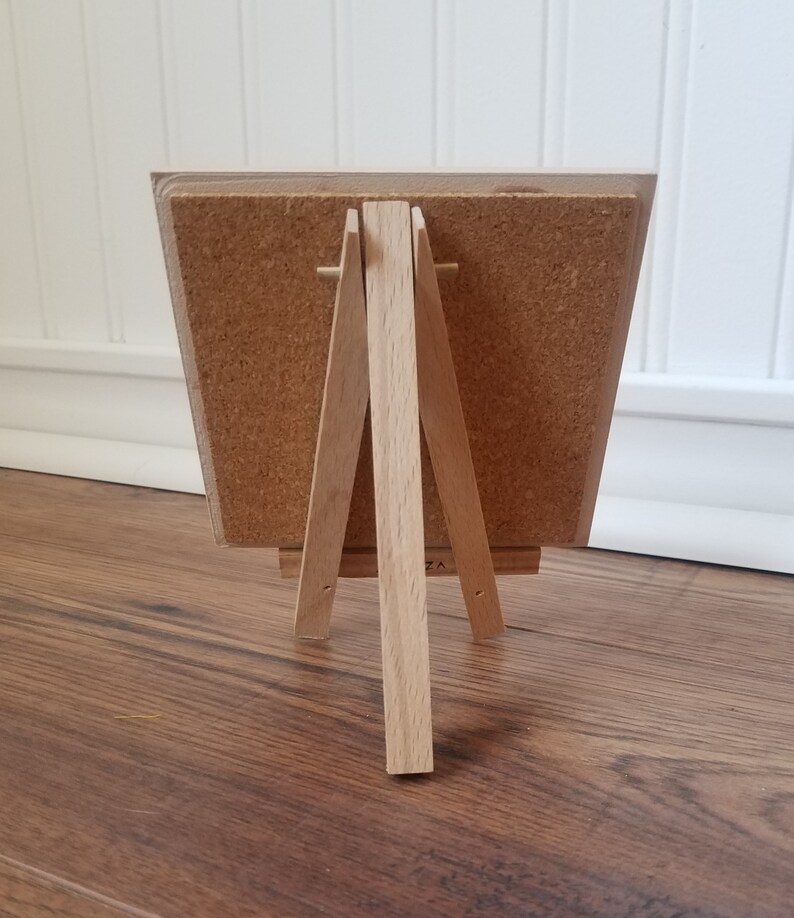 May include: A small wooden easel with a corkboard surface. The easel is made of natural wood and has a simple design.