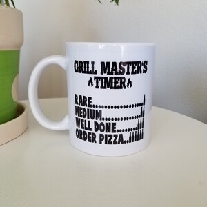 May include: White ceramic mug with a black graphic that reads "Grill Master's Timer" with flames on either side of the word "Timer". Below the text is a list of doneness levels for grilling meat: "Rare", "Medium", "Well Done", and "Order Pizza" with a corresponding number of beer bottles next to each level.