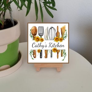 May include: A square sign with the text "Cathy's Kitchen" and illustrations of kitchen utensils, sunflowers, and greenery. The sign is displayed on a small wooden easel, with a potted plant in the background.