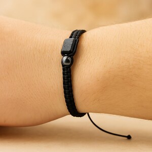 May include: Black braided bracelet with a square bead and a round bead. The bracelet is worn on a wrist and has a sliding knot closure. The beads are dark gray and the cord is black.