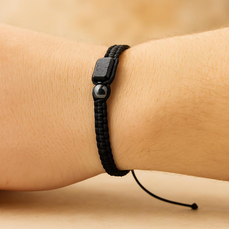 May include: Black braided bracelet with a square bead and a round bead. The bracelet is worn on a wrist and has a sliding knot closure. The beads are dark gray and the cord is black.