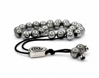 May include: A bracelet featuring a series of polished, metallic gray spherical beads strung on a black cord. The design includes a silver-toned metal charm with a spiral engraving and two smaller beads at the end of the adjustable cord closure.
