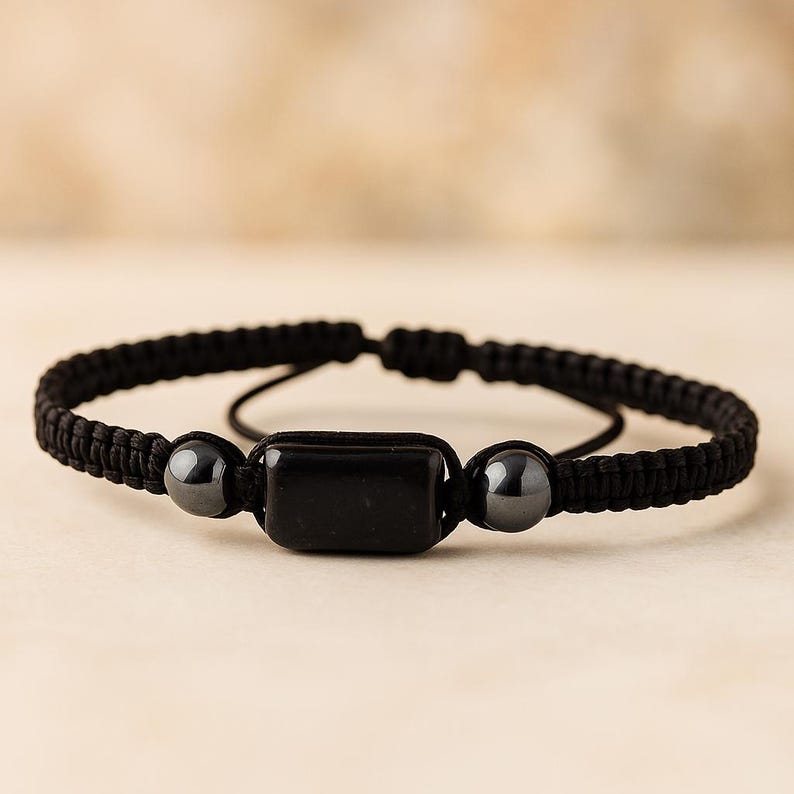 May include: Black braided bracelet featuring a rectangular black stone and two smaller, round black stones. The bracelet is adjustable and has a simple, minimalist design. This accessory is suitable for everyday wear.