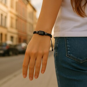 May include: Black bracelet featuring a rectangular black stone and two smaller, round, dark gray stones. The bracelet is made of a woven black cord and is adjustable. The bracelet is worn on a wrist.