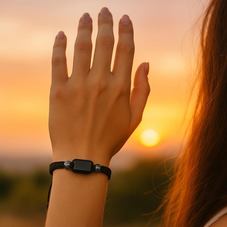 May include: Black bracelet with a rectangular black stone and two small silver beads. The bracelet is made of a woven black cord. The background is a sunset with a bright orange and yellow sky.